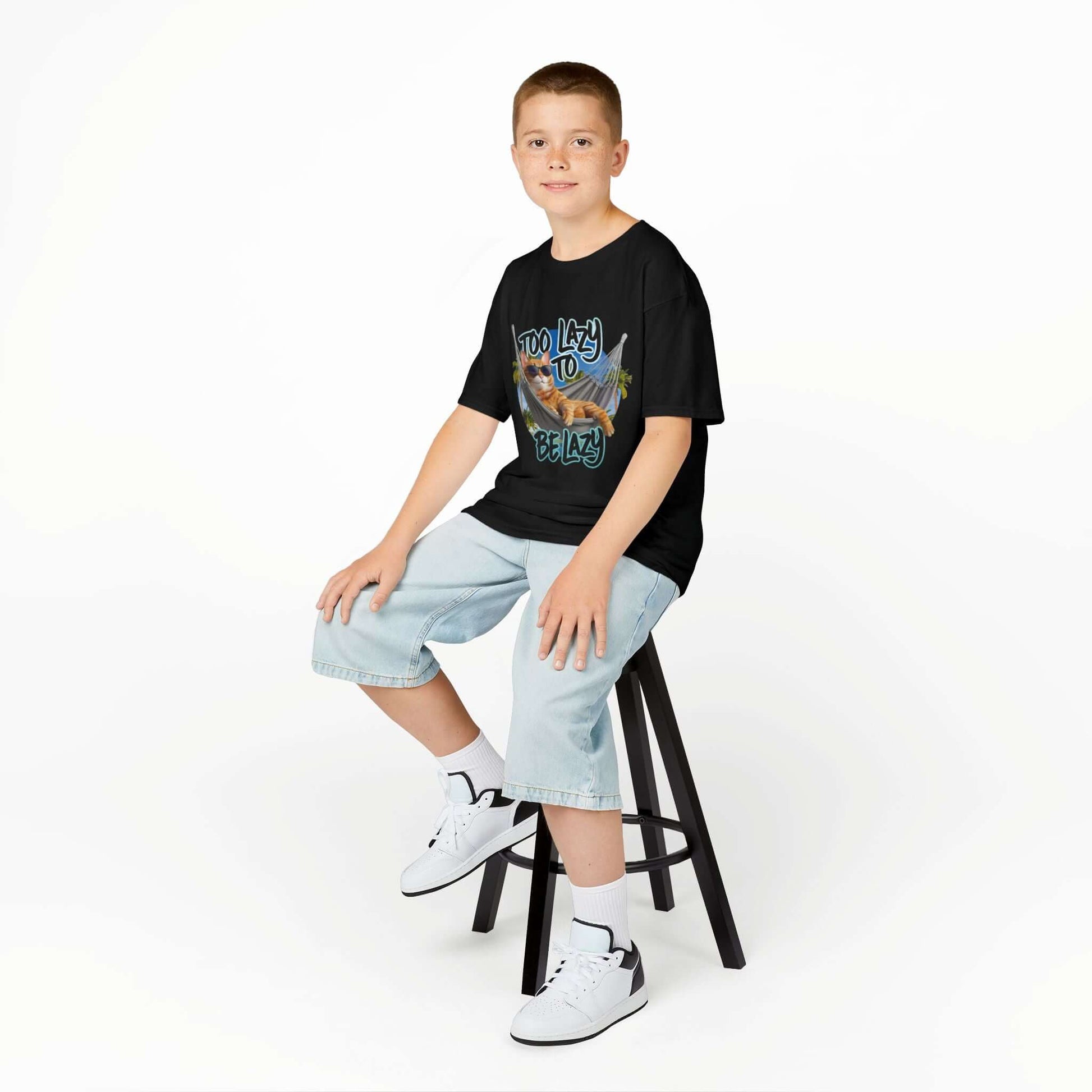 Boy sitting on a stool wearing a black kids tee with lazy cat at beach design and 'Too Lazy to be Lazy' text.