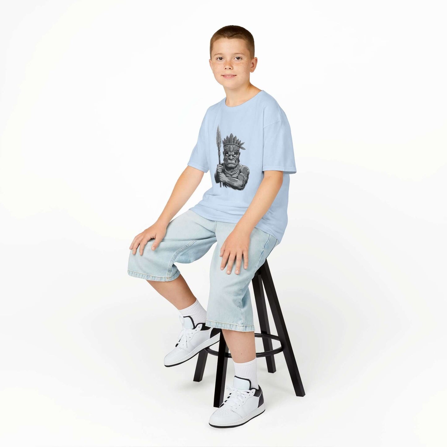 Boy wearing a blue kids tee with black and white Tiki character design, sitting on a stool, showcasing trendy fashion.
