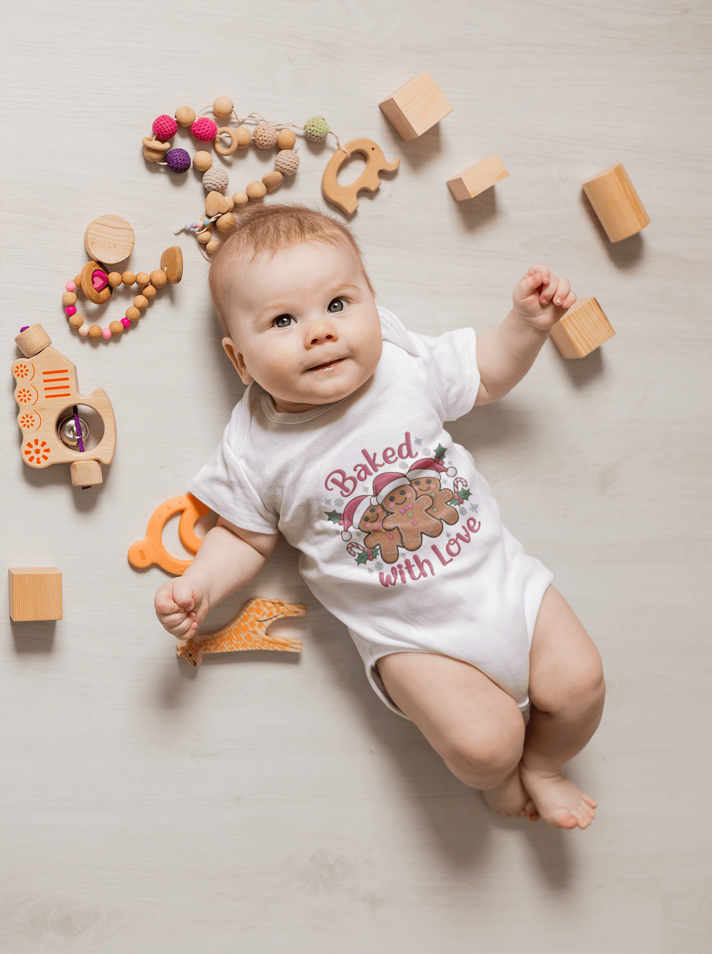 Baby in a festive white bodysuit with gingerbread men and 'Baked with Love' design, surrounded by playful toys on the floor.