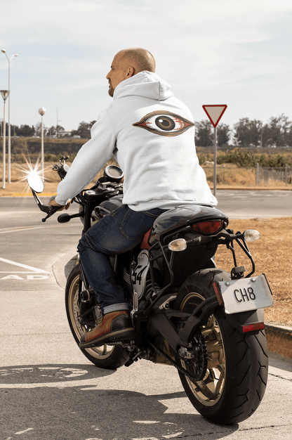 Man wearing a hoodie with a bloody brown eye design while riding a motorcycle on a sunny day.