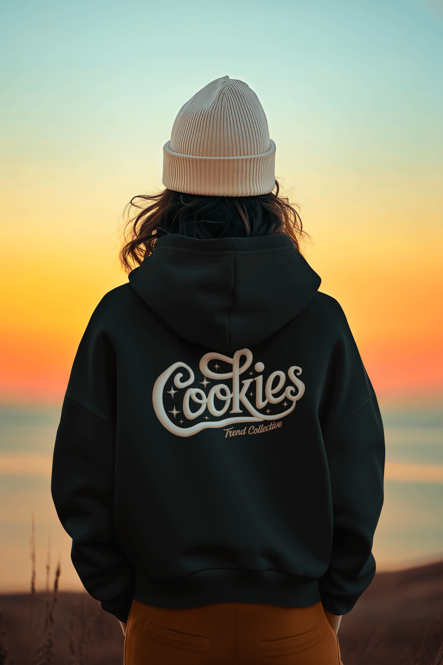 Back view of a person wearing a hooded sweatshirt with vintage 'Cookies Trend Collective' design against a sunset backdrop.