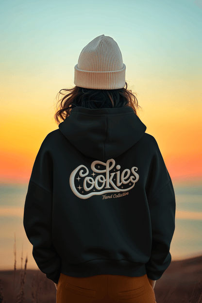 Back view of a person wearing a hooded sweatshirt with vintage 'Cookies Trend Collective' design against a sunset backdrop.