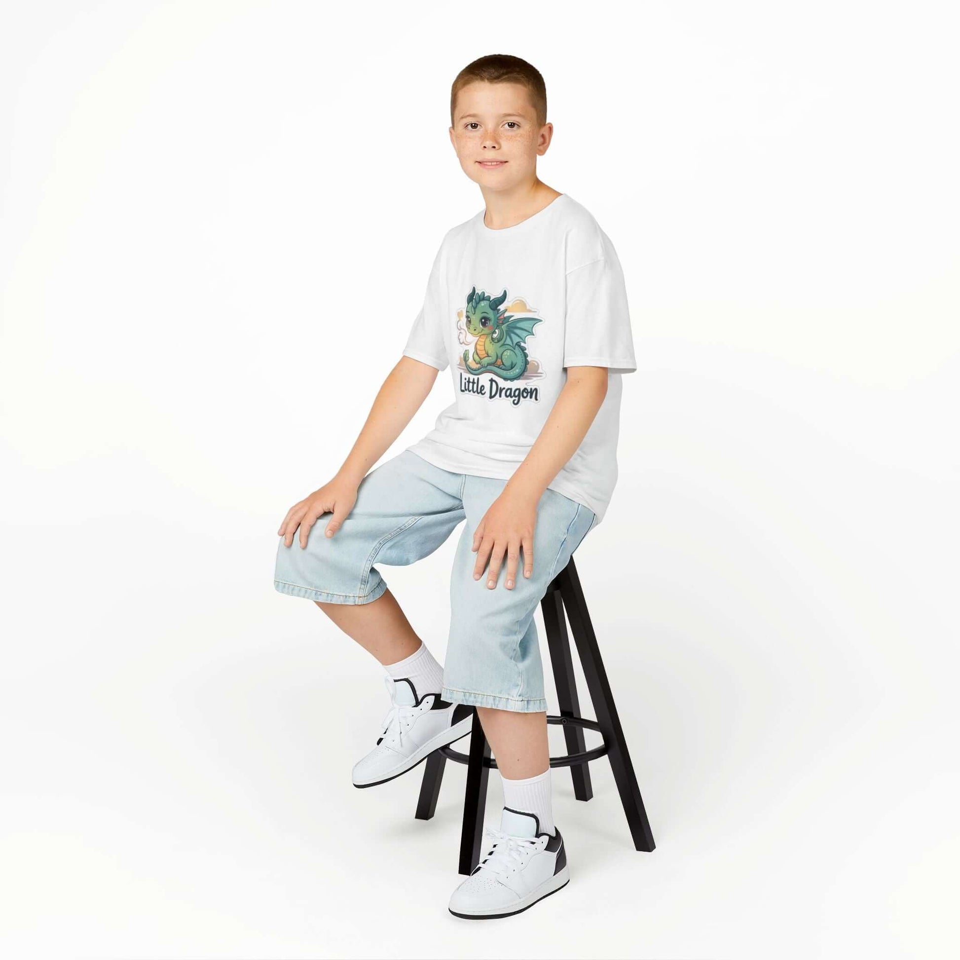 Boy wearing a 'Little Dragon' cartoon character Kids Tee, sitting on a stool, showcasing playful design.