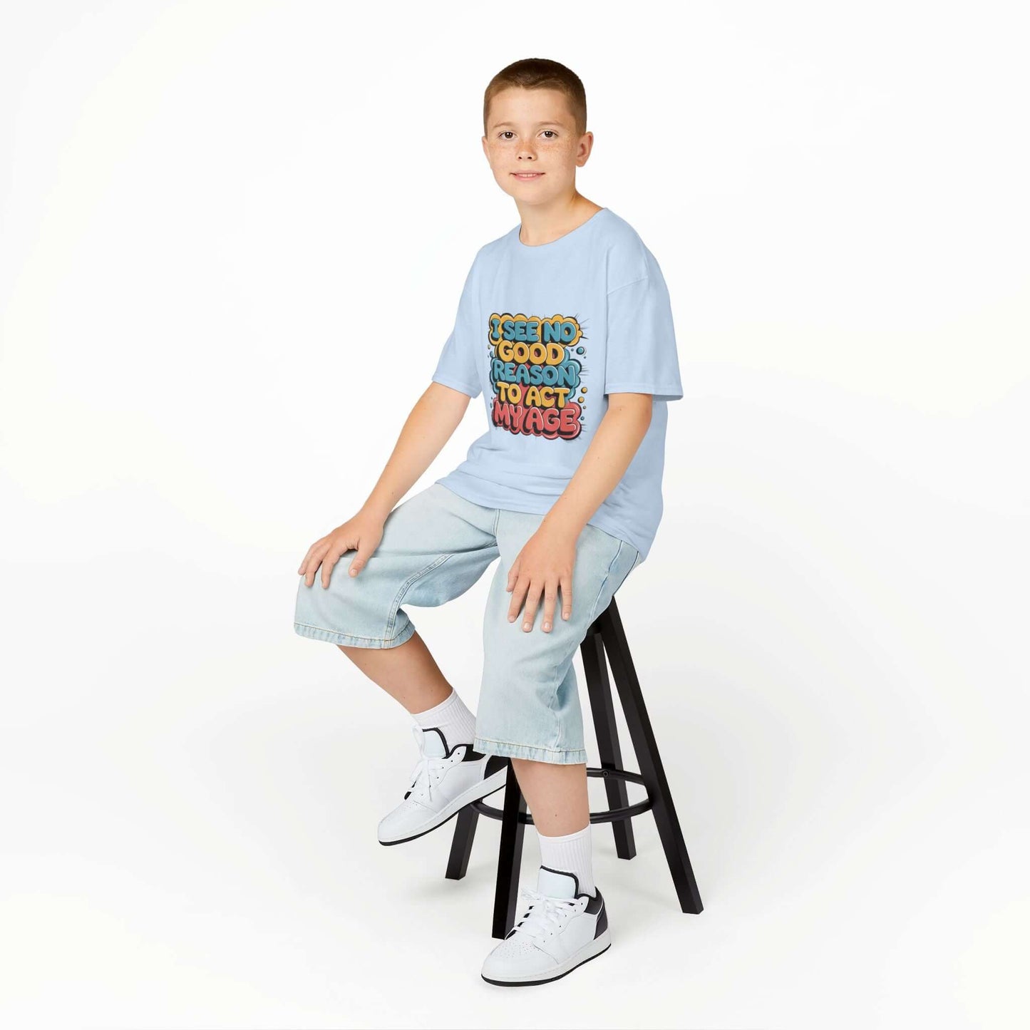 Kid sitting on stool wearing blue tee with colorful graffiti design and funny quote 'I see no good reason to act my age'.