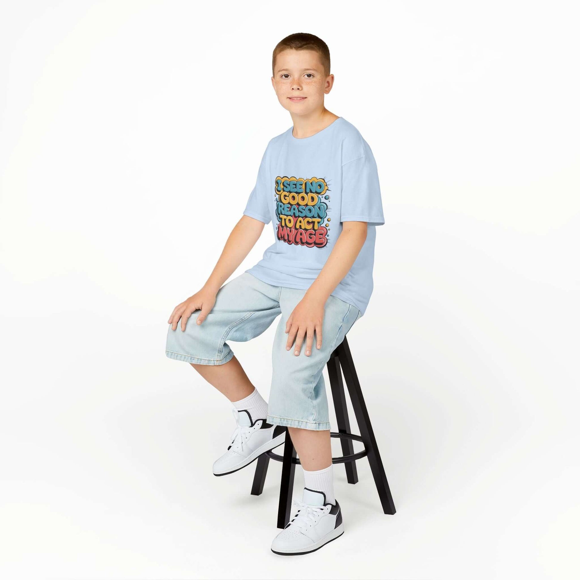 Kid sitting on stool wearing blue tee with colorful graffiti design and funny quote 'I see no good reason to act my age'.
